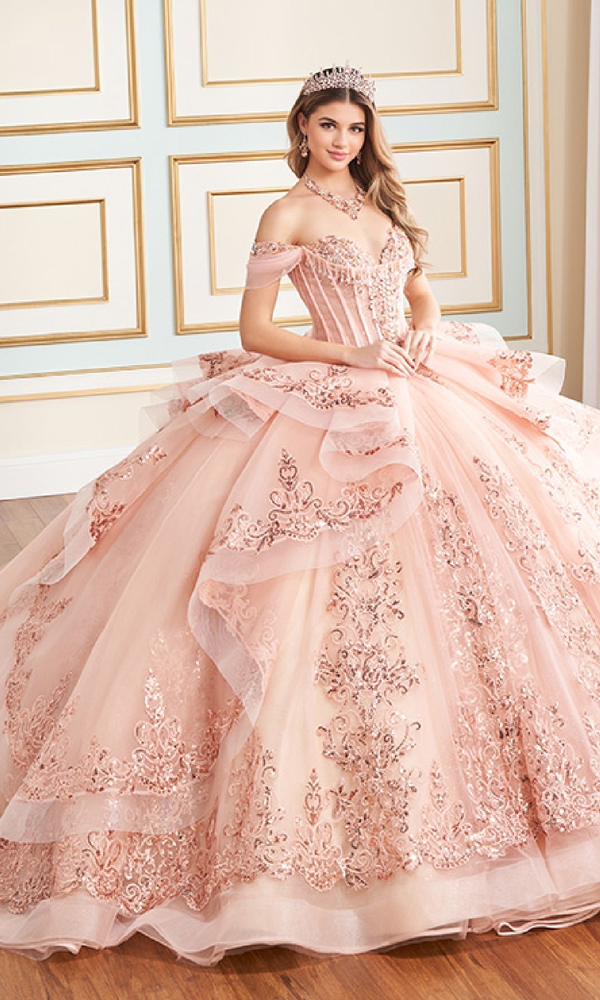 A woman stands indoors wearing the Quinceanera Dress PR30171 By Princesa, an elaborate off-the-shoulder pink gown with floral embroidery, glitter tulle, a tiara, and jewelry.
